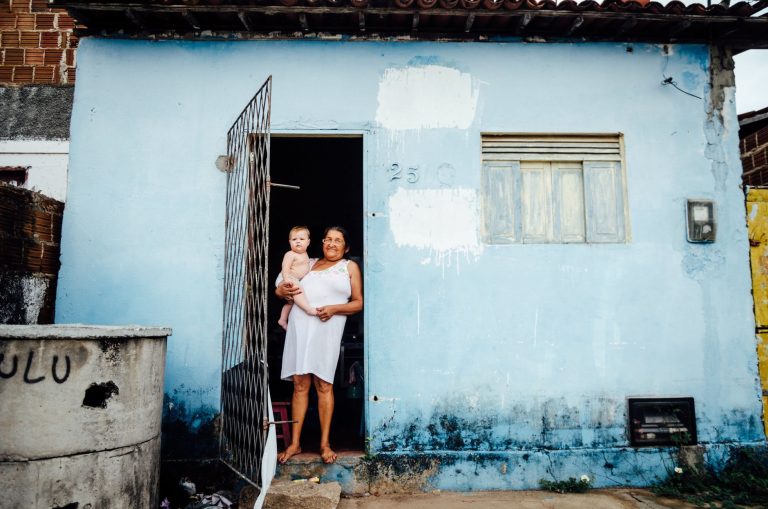 Moradora segurando um bebê na entrada de uma casa azul.