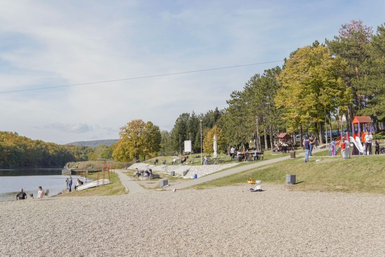 Guararema anuncia ampliação do Horto Parque Educacional com foco em turismo sustentável Caminho arborizado à margem de um rio, com árvores e um playground ao fundo, em um parque.