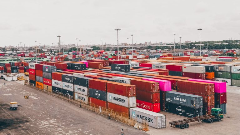 Contêineres coloridos empilhados em um porto movimentado, representando a logística do comércio internacional.